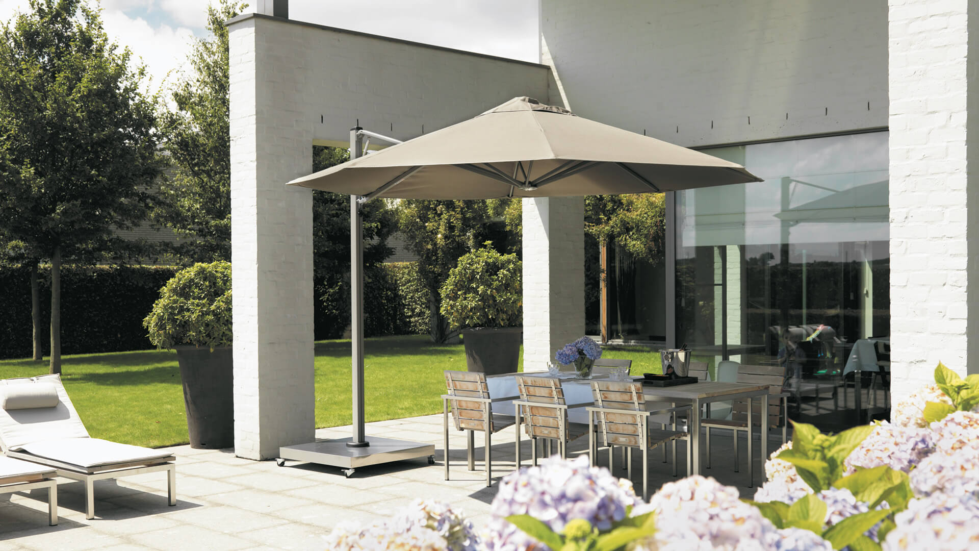 En terrasse med en parasol og havemøbler. Der er solskin, blomster der er sprunget ud og der står også to liggestole til venstre for terrassen.
